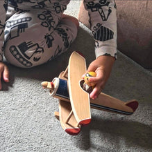 Load image into Gallery viewer, A child's hand pushing a wooden toy airplane with red and blue accents, with the child wearing gray and black construction-themed pajamas sitting on a gray carpet.